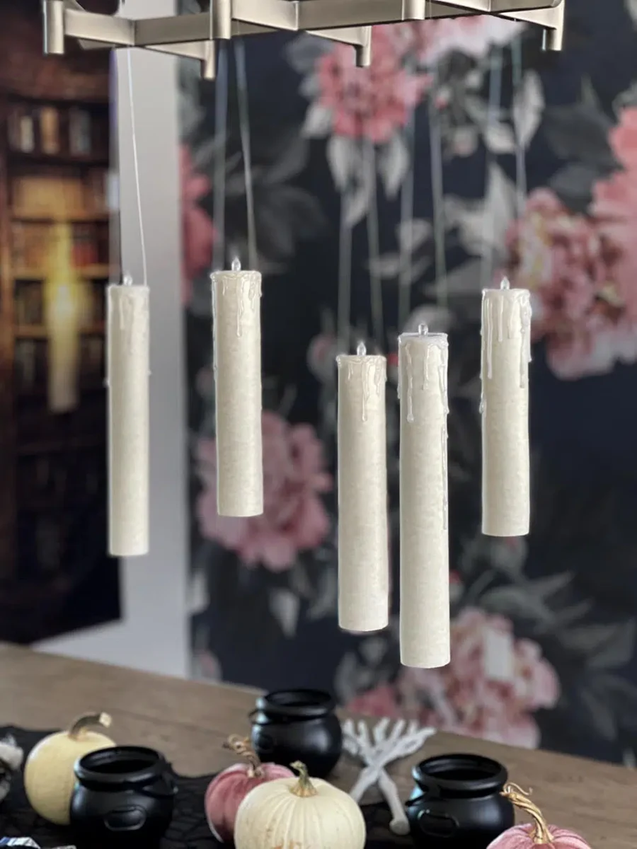 Five white candles hang from nearly invisible strings, appearing to float in mid-air. Below them are small cauldrons, white and pink pumpkins, and a wooden table. The background features floral wallpaper and a bookshelf.