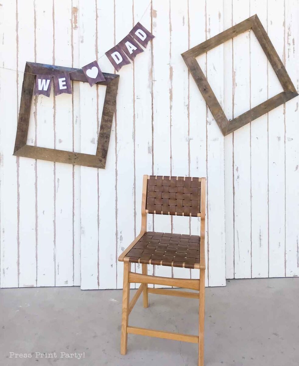 A wooden chair sits in front of a white, rustic plank wall with two empty wooden frames and a cheerful Fathers Day banner reading "WE ♥ DAD" hanging between them.