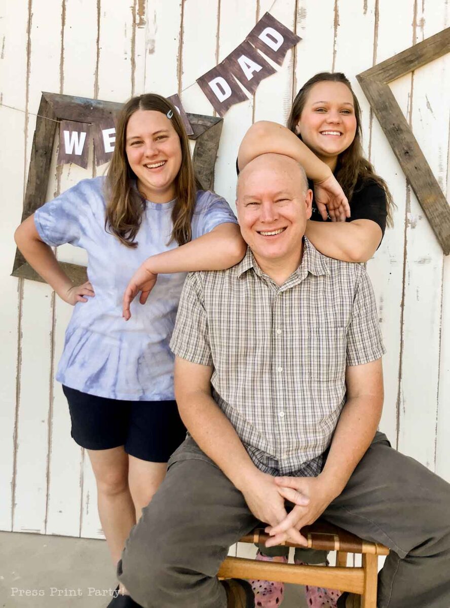 A smiling man sits on a stool with two smiling women standing behind him, one resting her arm on his shoulders. A rustic white wall in the background displays a festive Fathers Day banner that reads "WE ♥ DAD.