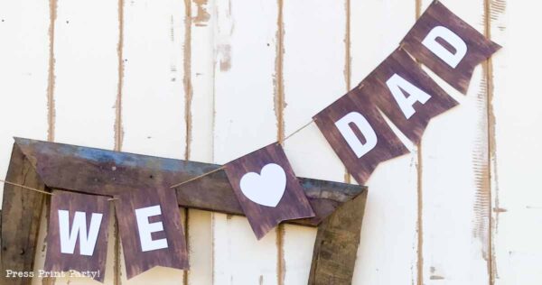 A rustic fathers day banner with dark brown flags reads "WE ♥ DAD" in white letters, hanging against a light-colored wooden wall.