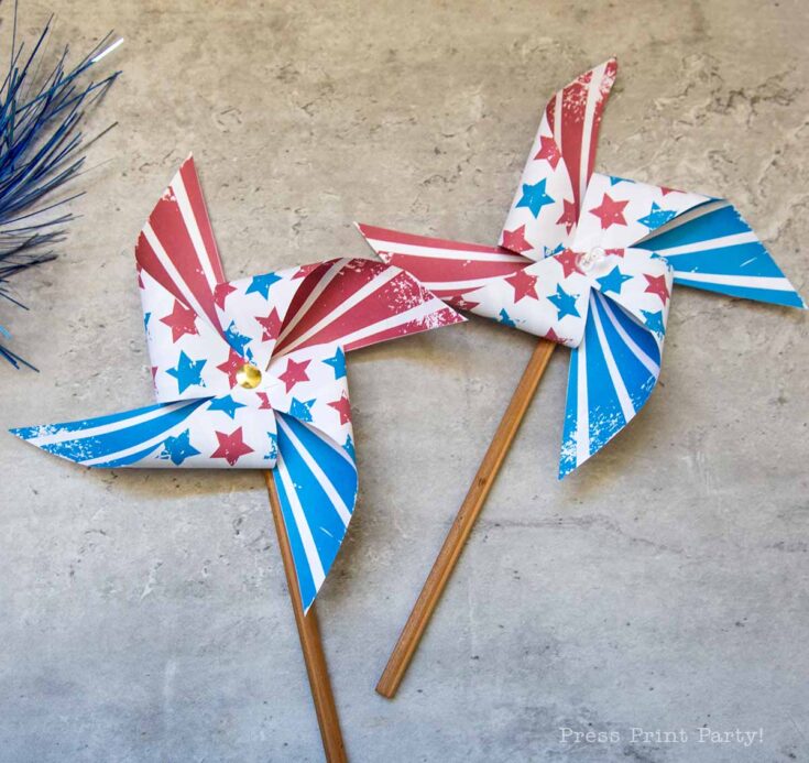 Two patriotic pinwheels with red, white, and blue star and stripe patterns on wooden sticks lie on a gray surface, evoking a festive theme. Blue tinsel is partially visible in the corner.