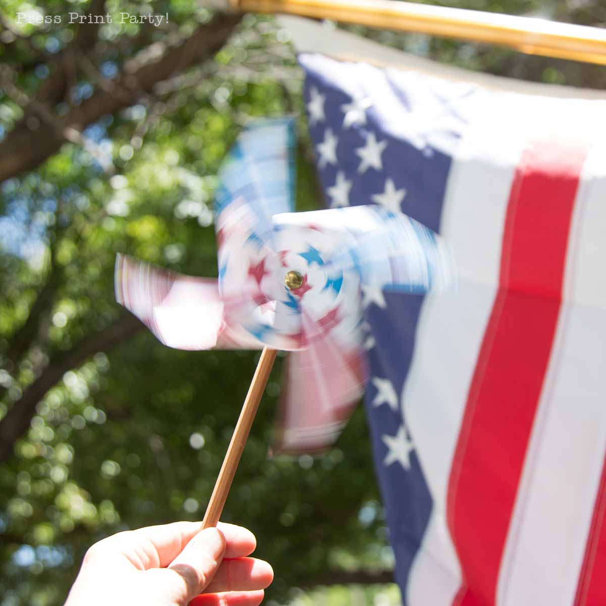 A hand holds a spinning patriotic pinwheel in red, white, and blue in front of a blurred American flag outdoors, with green trees in the background.