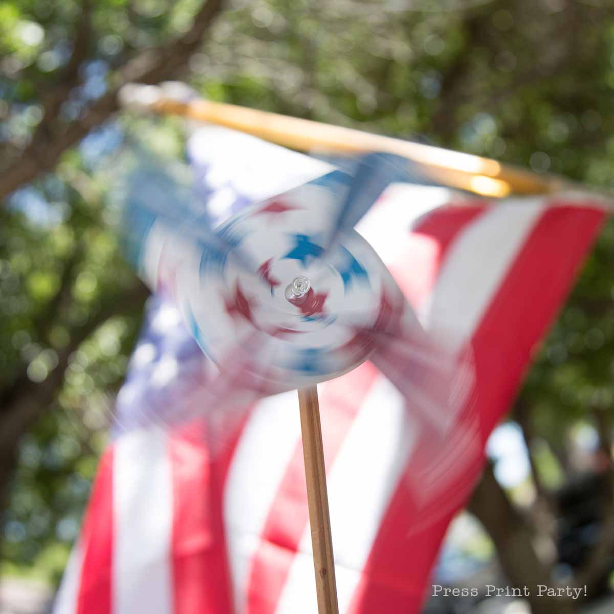 A patriotic pinwheel with red and blue stars spins in front of a blurred American flag outdoors, framed by green trees in the background.