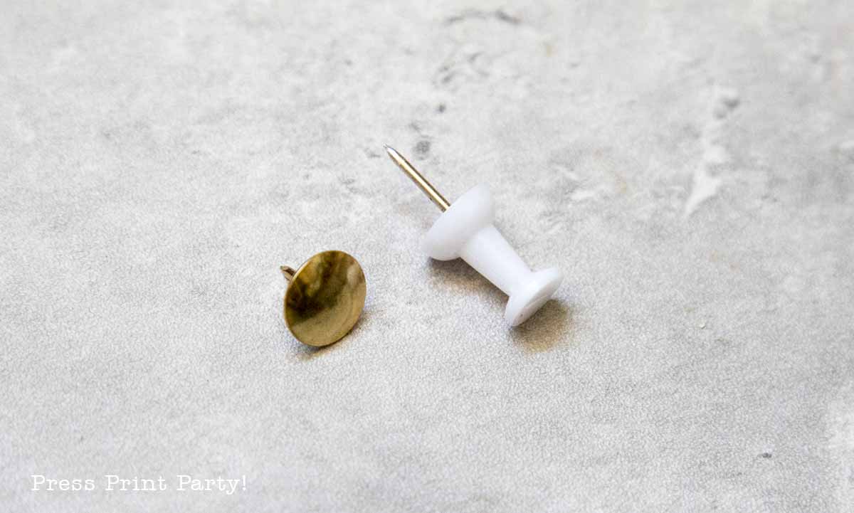 A gold brad and a white push pin, perfect for securing patriotic pinwheels, lie on a light gray, textured surface. The brad is flat and round, while the push pin stands upright with a cylindrical shape.