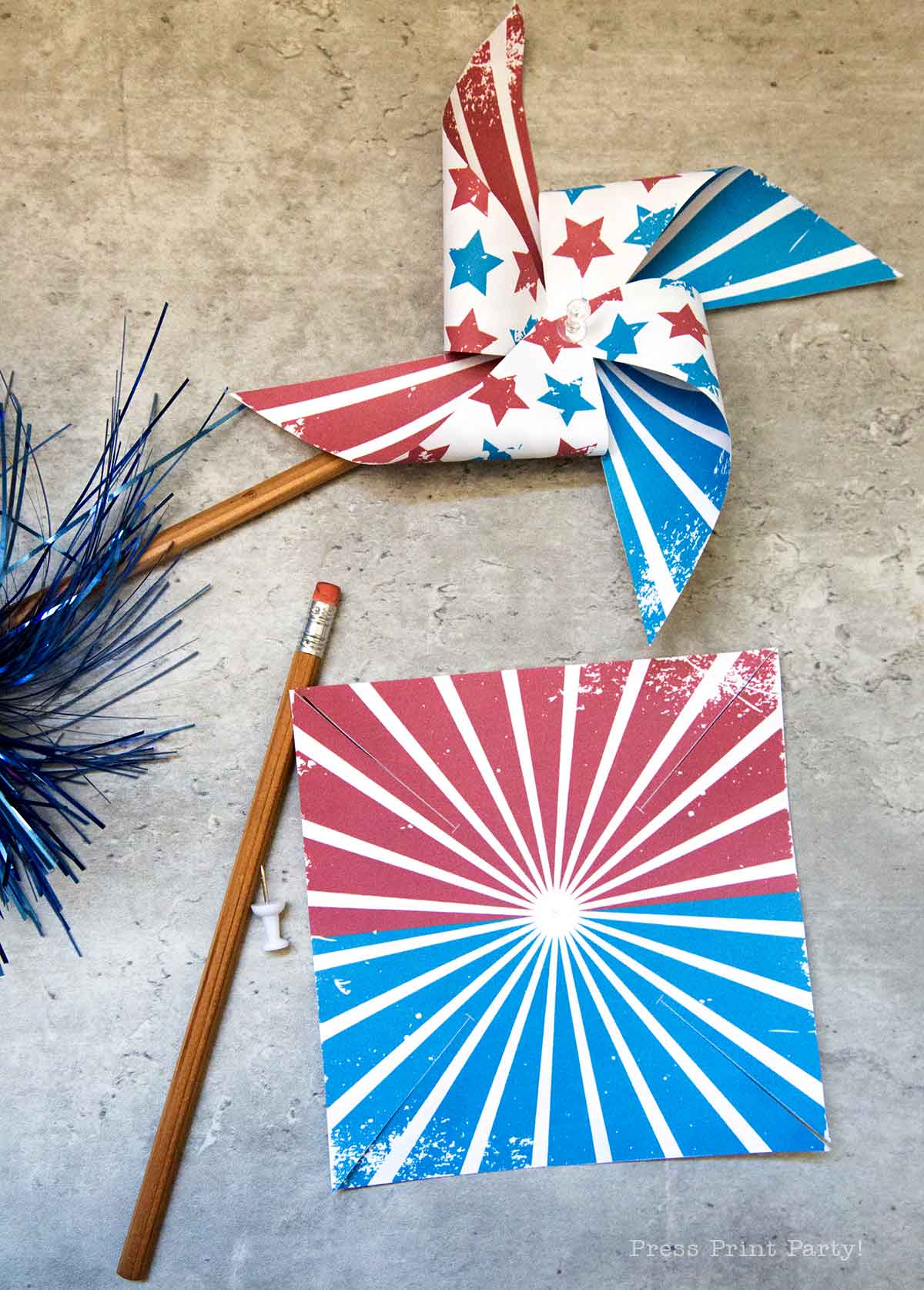 A red, white, and blue patriotic pinwheel with star patterns is attached to a pencil. Nearby is a matching square paper with a sunburst design, a push pin, another pencil, and blue metallic tinsel on a gray surface.