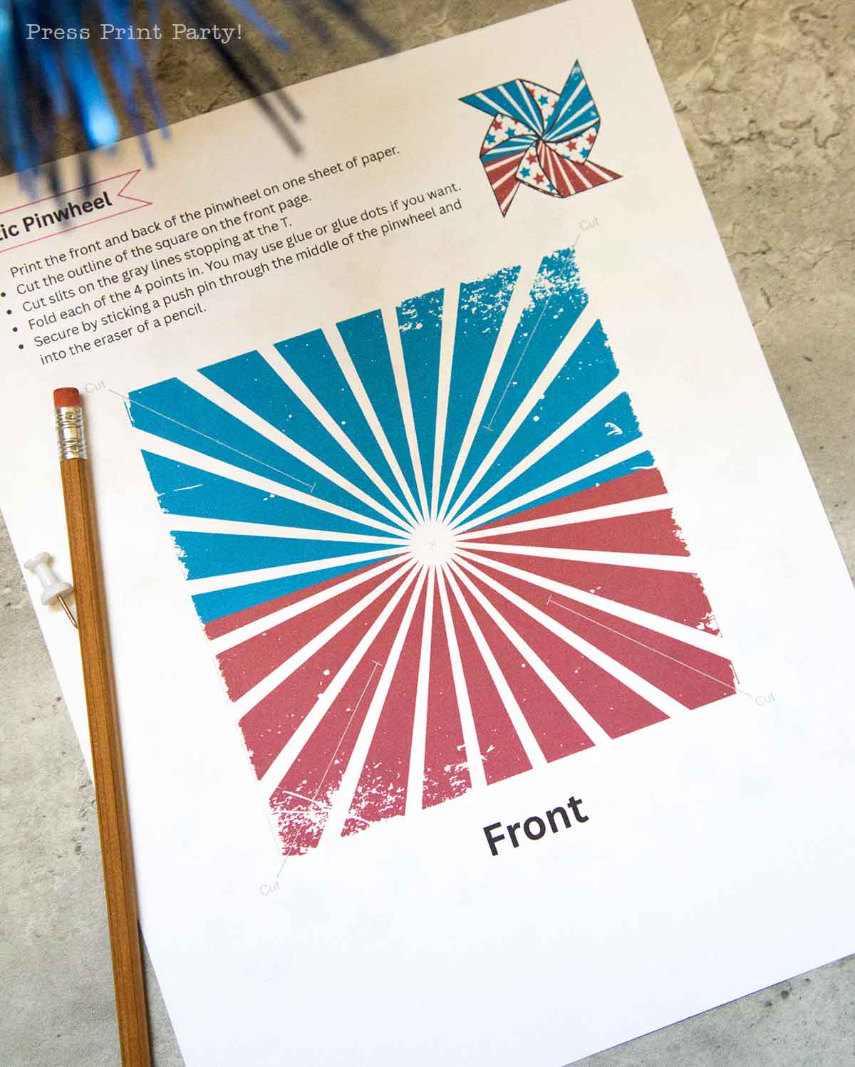 A printed pinwheel template labeled "Front" features a red, blue, and white starburst pattern—perfect for creating patriotic pinwheels. A pencil and push pin lie beside the sheet on a marble surface, with assembly instructions at the top.