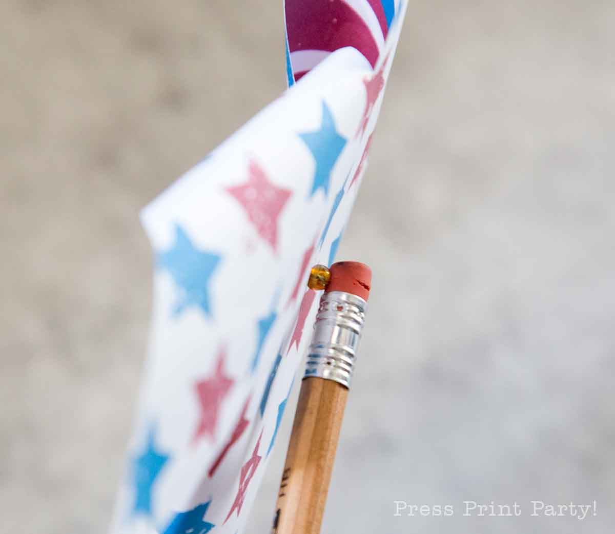A close-up of a pencil holding a patriotic pinwheel made of paper decorated with red and blue stars. The background is blurred, and "Press Print Party!" is faintly visible in the lower right corner.