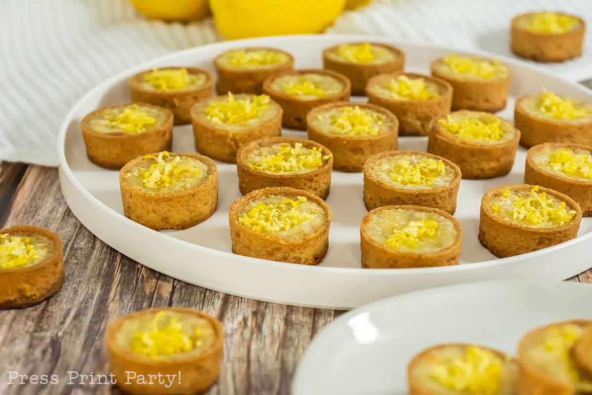 A white tray filled with small, round lemon tarts from a classic mini lemon tarts recipe sits on a wooden table. More tarts are on a plate in the foreground, with whole lemons displayed in the background.