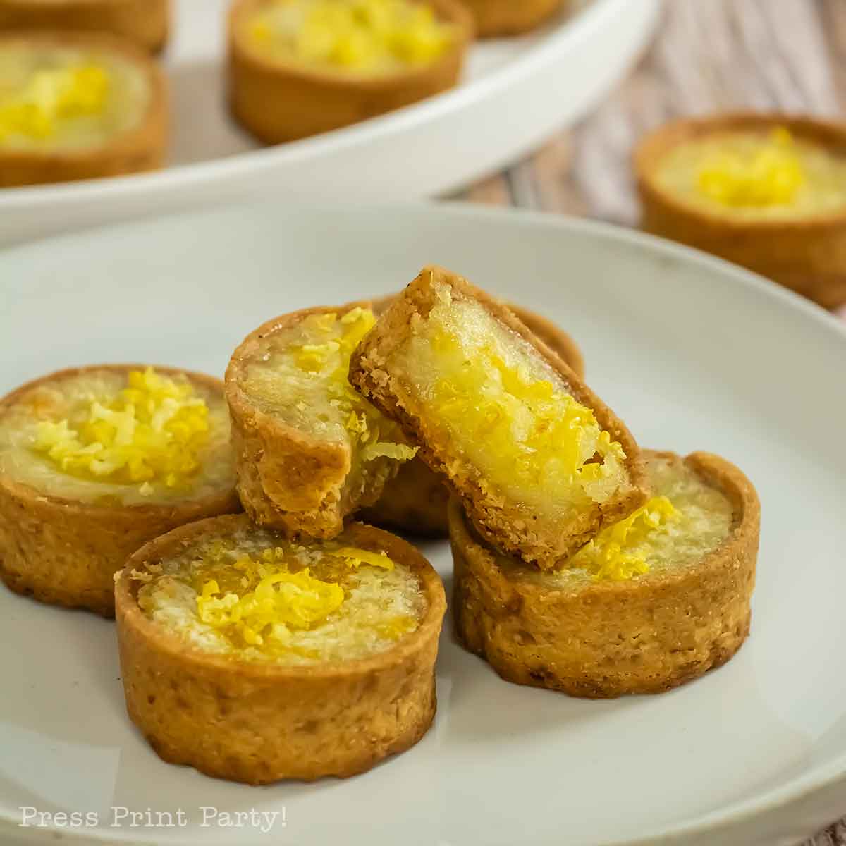 A plate of round mini lemon tarts with golden crusts and a yellow filling, topped with lemon zest. One tart is cut in half, showing the moist interior. More from this mini lemon tarts recipe are on a tray in the background.