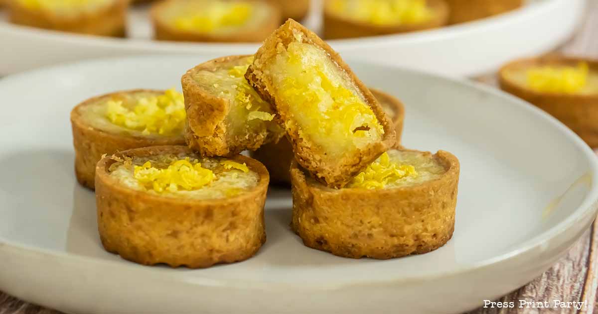 A plate with several small lemon tarts featuring golden brown crusts and yellow lemon filling, perfect for any mini lemon tarts recipe, with one tart cut in half and stacked on top. Other tarts are visible in the background on another plate.