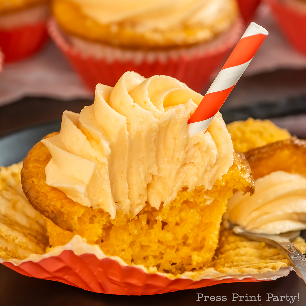 Close-up of a cupcake with swirled yellow frosting, partially unwrapped and bitten into, with a red-and-white striped straw inserted on top. Other frosted cupcakes are visible in the background.