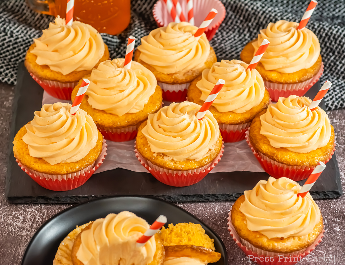 A group of cupcakes with swirled orange frosting and red-striped straws, each in a red paper liner, displayed on a tray with a textured background. One cupcake is cut open to show the yellow cake inside.