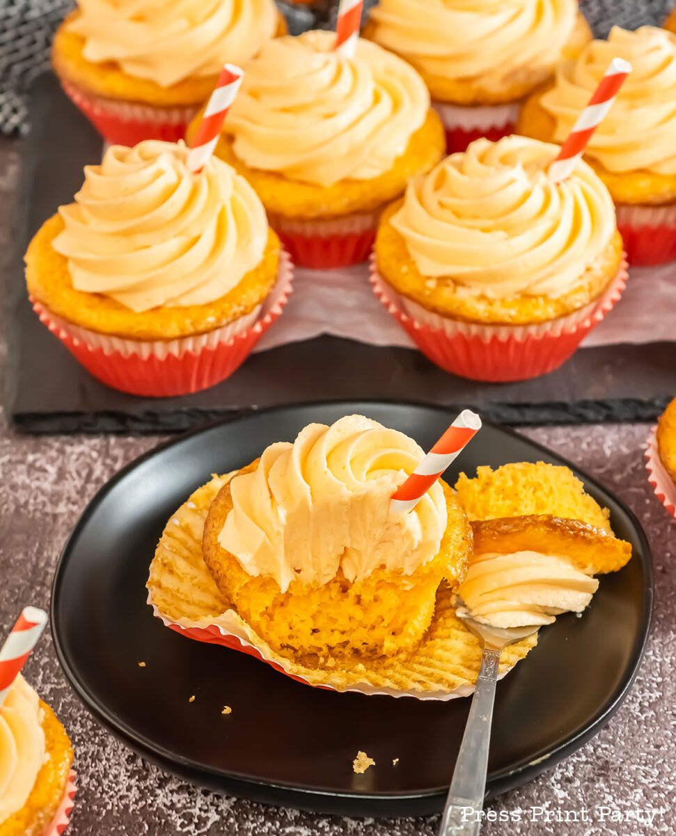 A plate with a frosted cupcake, partially unwrapped and cut open, is in front of several more cupcakes topped with swirled frosting and red-striped straws, all in red liners on a dark surface.