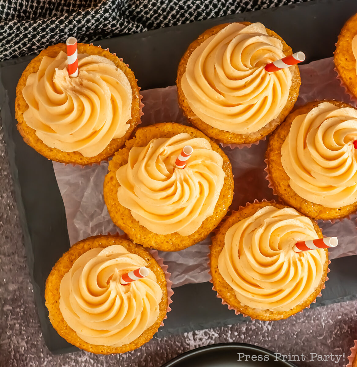 Six cupcakes with swirled orange frosting and red-striped straws sit on parchment paper atop a dark tray, viewed from above. A cloth napkin is partially visible in the corner.