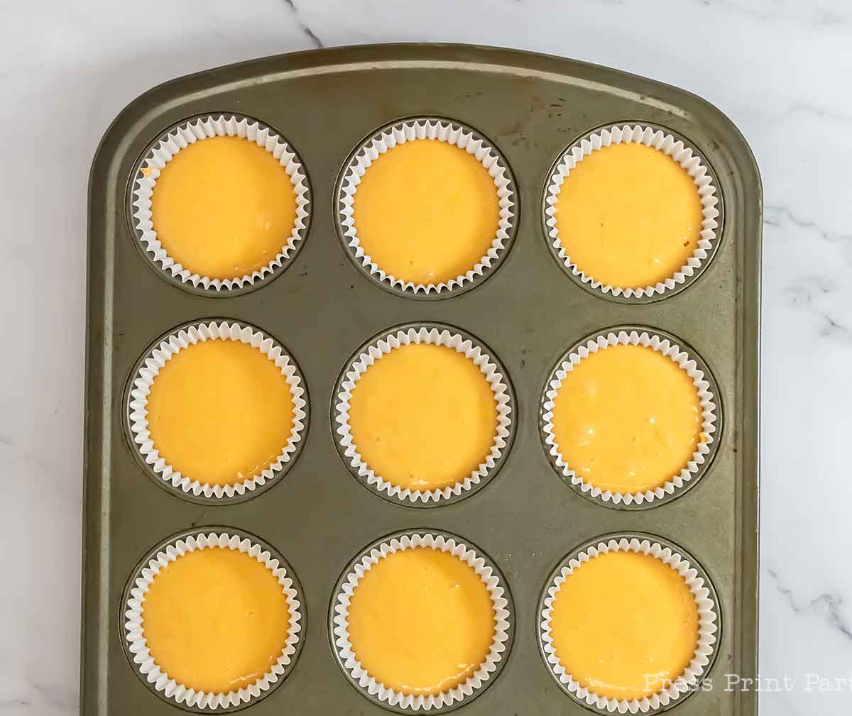 A muffin tin filled with nine paper cupcake liners, each holding yellow batter, sits on a white marble surface.