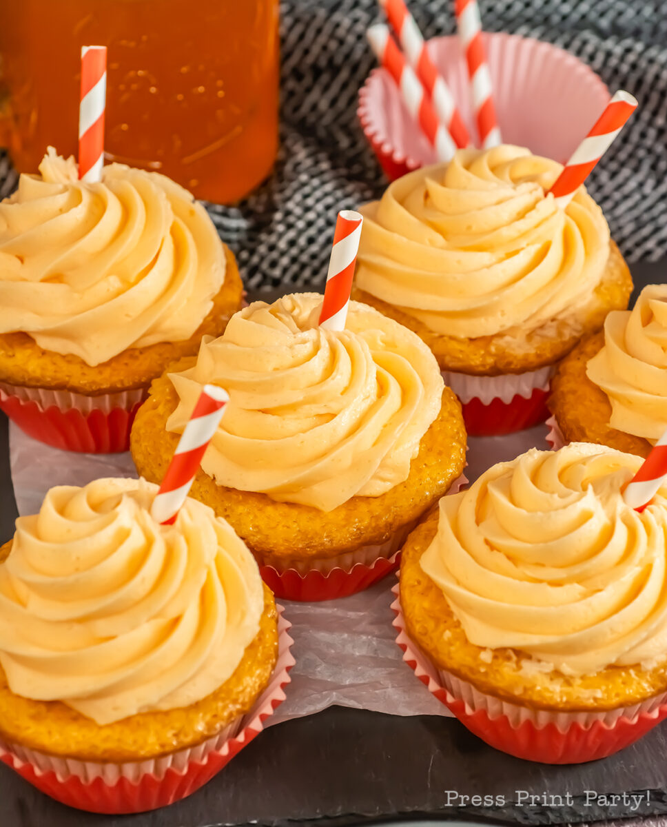 Six cupcakes with orange frosting are topped with red and white striped paper straws. The cupcakes are in red paper liners and arranged on a tray, with a jar of orange liquid blurred in the background.
