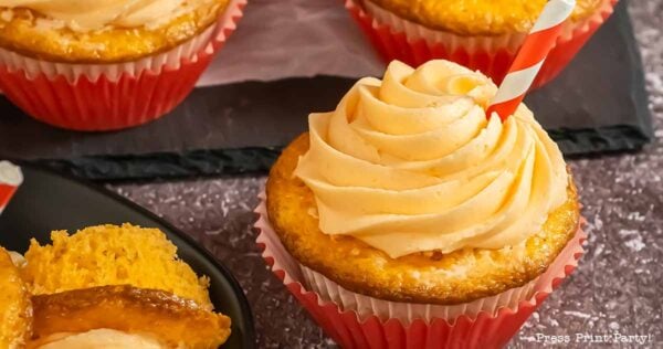 Cupcakes with orange frosting in red and white liners are displayed on a dark surface. One cupcake is topped with a red and white striped straw, and another cupcake is partially cut open, revealing its moist interior.