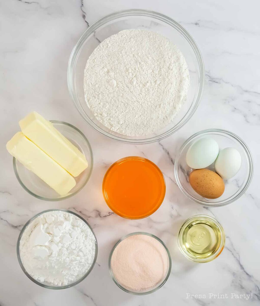 Bowls containing flour, powdered sugar, orange gelatin powder, oil, three eggs, and two sticks of butter arranged neatly on a white marble surface.