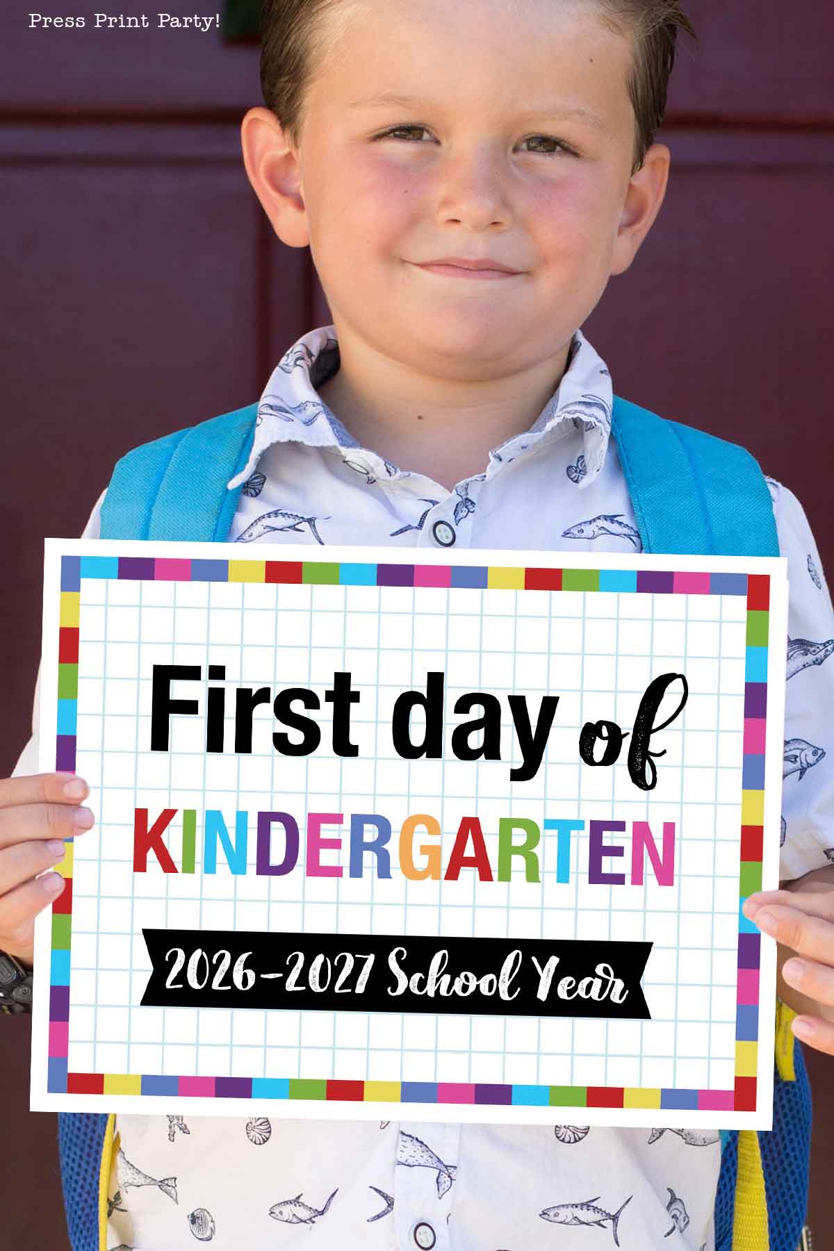 Smiling young boy with a blue backpack holds a colorful first day of school printable sign that reads, "First day of Kindergarten, 2026-2027 School Year," standing in front of a maroon background.