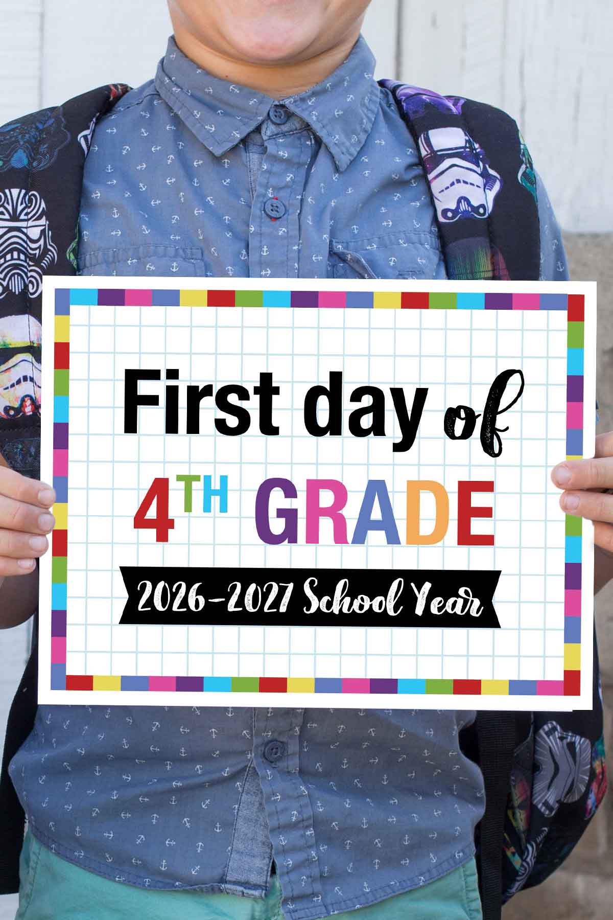 A child in a blue patterned shirt holds a vibrant first day of school printable sign reading "First day of 4th grade, 2026-2027 school year" against a grid paper background. The child’s face is partially out of frame.