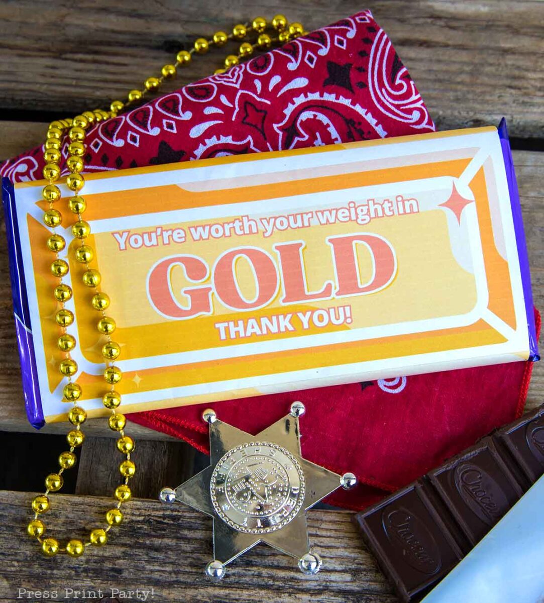 A perfect volunteer appreciation gift: A chocolate bar wrapped with "You're worth your weight in GOLD THANK YOU!" rests on a red bandana, gold bead necklace, and silver sheriff badge atop rustic wooden planks.