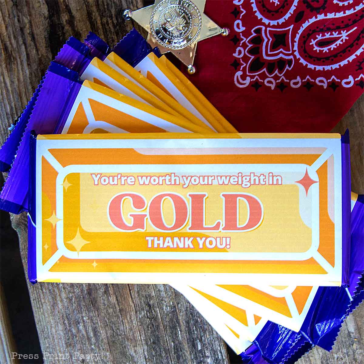 A stack of chocolate bars with gold and orange wrappers reads, "You're worth your weight in GOLD THANK YOU!" Nearby are a toy sheriff badge and a red bandana on a wooden surface—perfect as a volunteer appreciation gift.