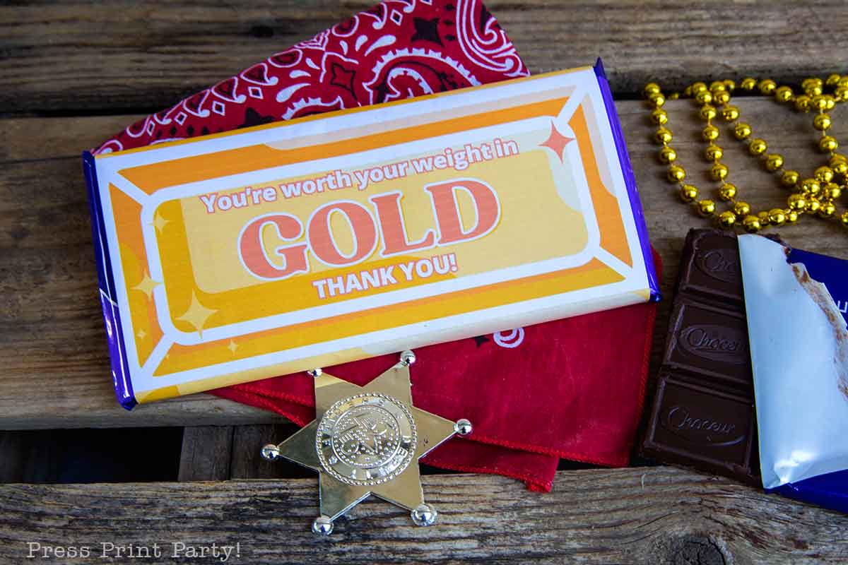 A chocolate bar with a wrapper reading "You're worth your weight in GOLD THANK YOU!" sits on a wooden surface next to a gold sheriff badge, red bandana, and gold beads—an ideal volunteer appreciation gift with part of the chocolate unwrapped.