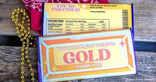 Two colorful candy bars on a wooden surface make the perfect volunteer appreciation gift. One wrapper says "YOU'RE PRECIOUS!" and the other features "You're worth your weight in GOLD! THANK YOU!" with a fun nutrition facts parody. Red bandana and gold beads nearby.