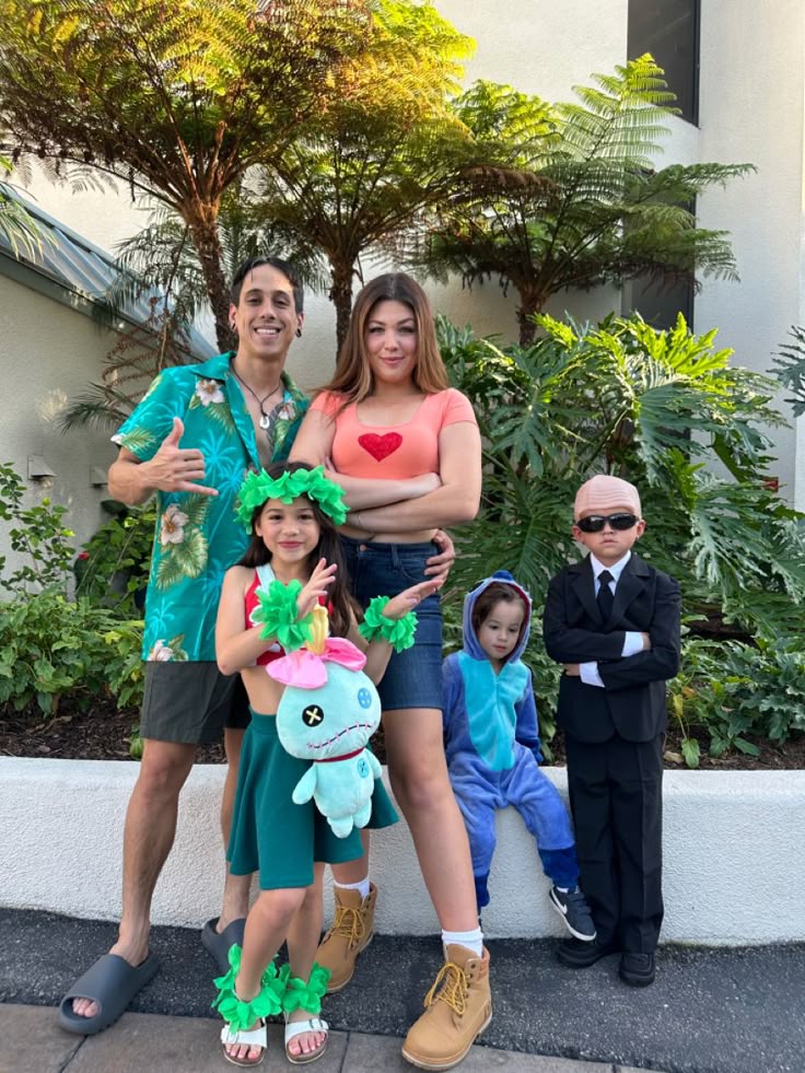 A family of five poses outdoors in costumes: a man in a floral shirt, a woman in a red heart shirt, a girl dressed as a leafy character holding a plush toy, a child in a blue animal suit, and a boy in a suit with sunglasses.