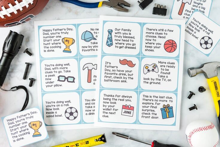 Two printed Father’s Day scavenger hunt clue cards are surrounded by tools, a football, a flashlight, a whistle, and other small items on a marble surface. The cards feature playful illustrations and text clues.