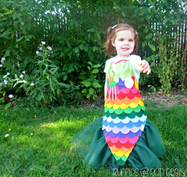 A young girl stands on grass, smiling and wearing a colorful homemade mermaid costume with rainbow scales and green fins. Lush green bushes and sunlight are in the background.