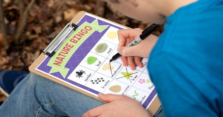 A person wearing a blue shirt holds a clipboard with a "Nature Bingo" sheet and marks off items like leaves and mushrooms while sitting outdoors, surrounded by fallen leaves.