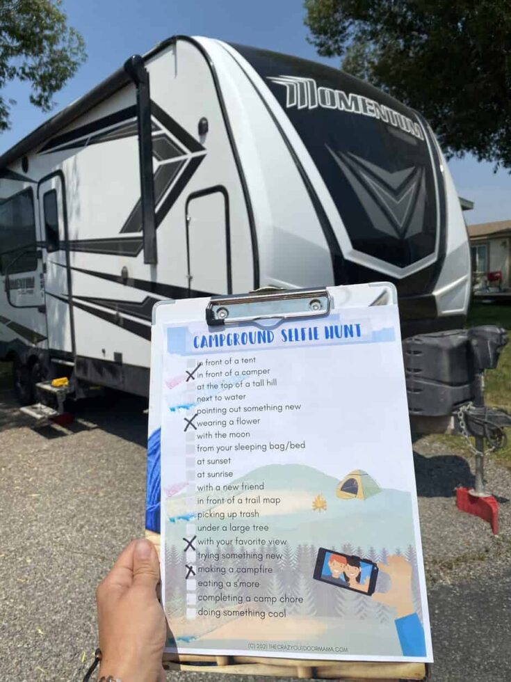 A hand holds a clipboard with a "Campground Selfie Hunt" checklist in front of a large white and black RV. Several items on the checklist are marked off. Trees and grass are visible in the background.