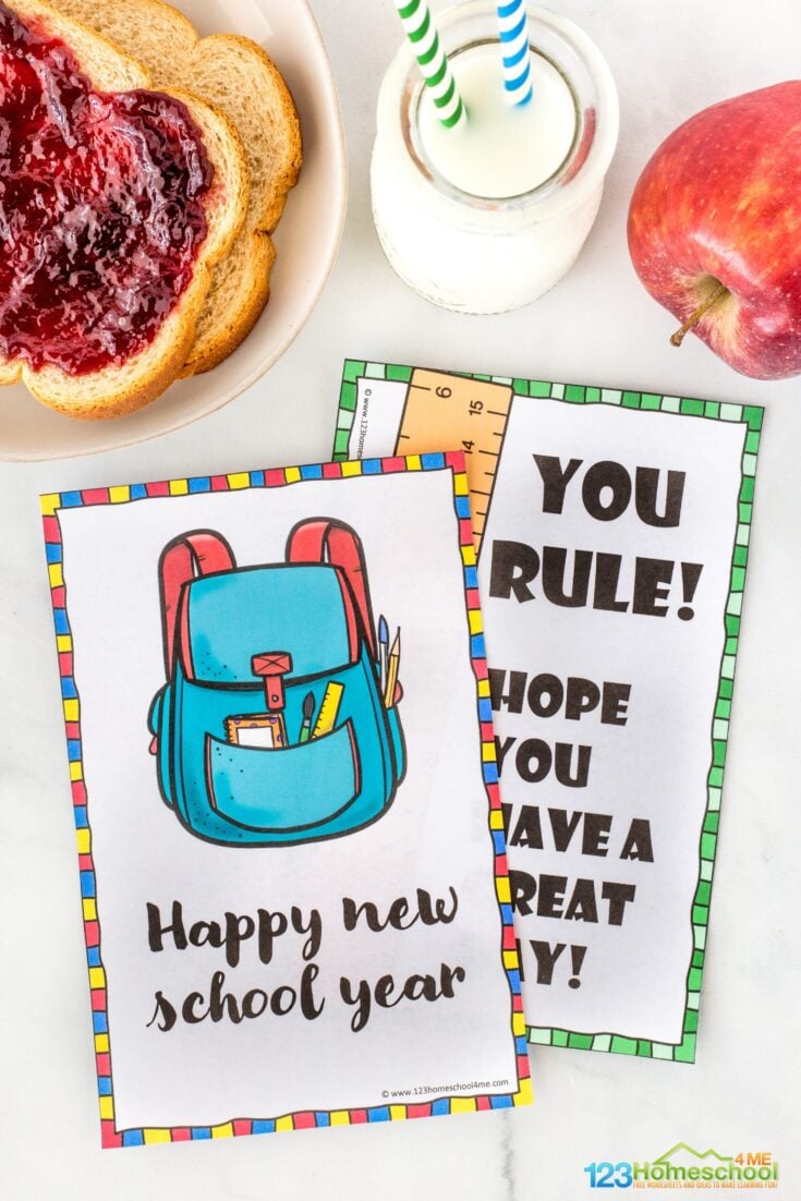 A card with a drawing of a backpack and school supplies says "Happy new school year." Next to it, another card reads "YOU RULE! HOPE YOU HAVE A GREAT DAY!" Jam toast, a glass of milk, and an apple are nearby.