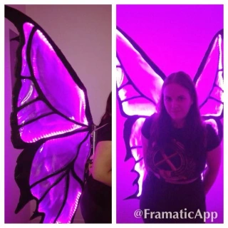 A woman wearing large glowing purple butterfly wings, shown from the side and from the back, standing indoors. The wings are illuminated with bright lights. "@FramaticApp" is in the corner.