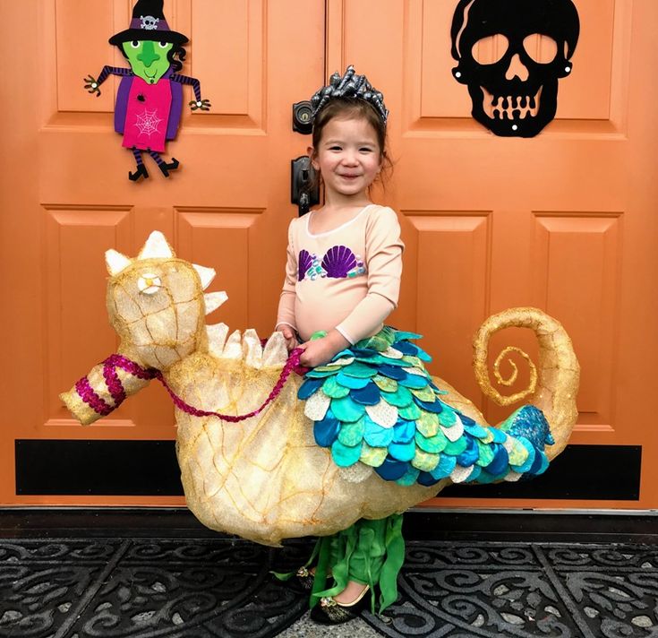 Young child in a mermaid costume with a colorful fish tail and seashell top, standing inside a large, golden seahorse costume. Halloween decorations, including a witch and skull, hang on an orange door behind her.