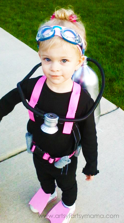 A young child wears a black outfit with pink accessories, flippers, a scuba tank, goggles, and a breathing tube—dressed as a scuba diver—standing on the sidewalk. Perfect for anyone searching for creative under the sea costume ideas.