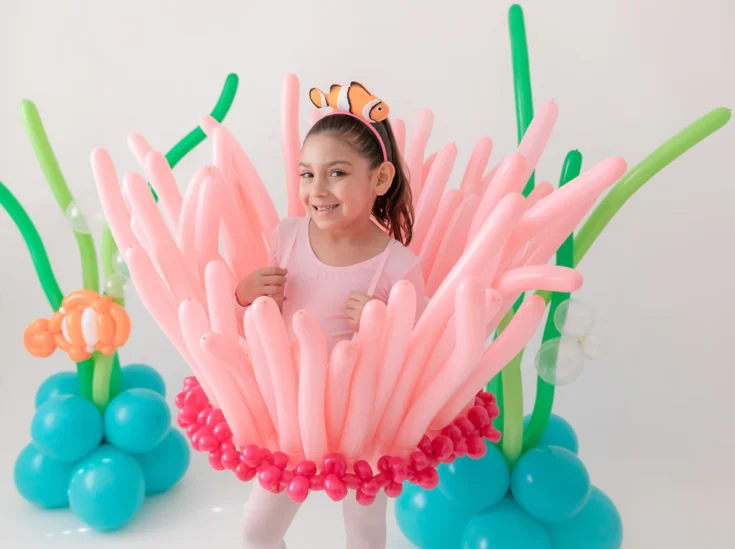 A smiling girl wearing a headband with a clownfish stands inside a costume made of pink balloons resembling sea anemone tentacles—one of the most creative under the sea costume ideas—surrounded by balloon decorations like underwater plants.