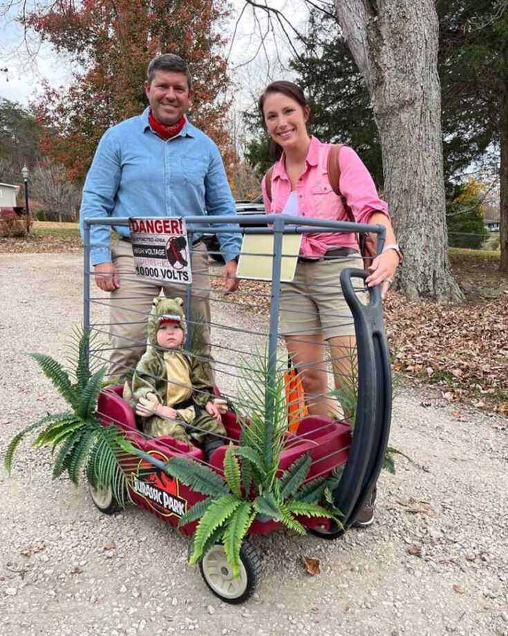A creative family costume idea: A man and woman dressed as Jurassic Park workers stand behind a red wagon decorated with plants and a “Danger: 10,000 Volts” sign, carrying their child in a dinosaur costume outdoors on a gravel path.
