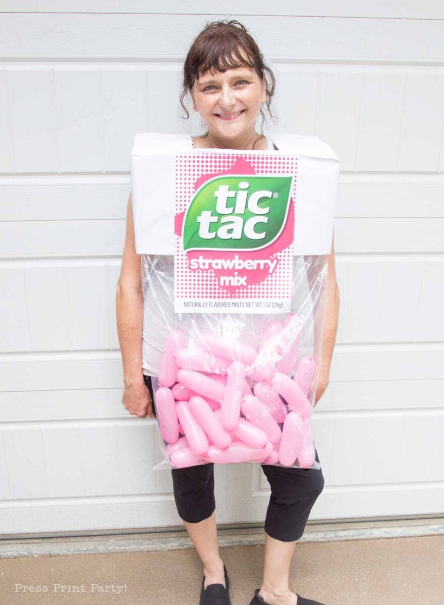 A woman stands in front of a garage door wearing a creative costume resembling a box of Strawberry Mix Tic Tacs made from cardboard and clear plastic, filled with large pink capsules.