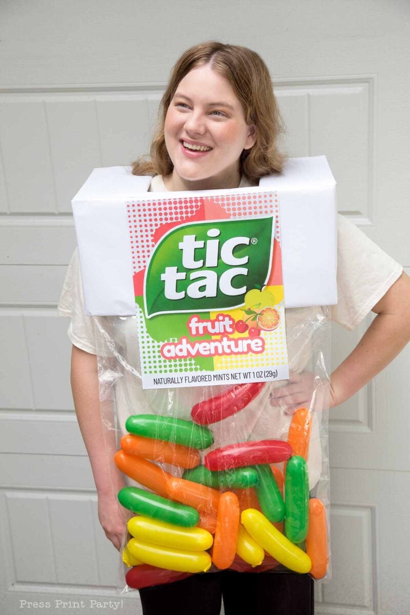 A person smiles while wearing a homemade Tic Tac Fruit Adventure costume made from a large plastic bag and a cardboard box with the Tic Tac label, filled with oversized colorful capsules.