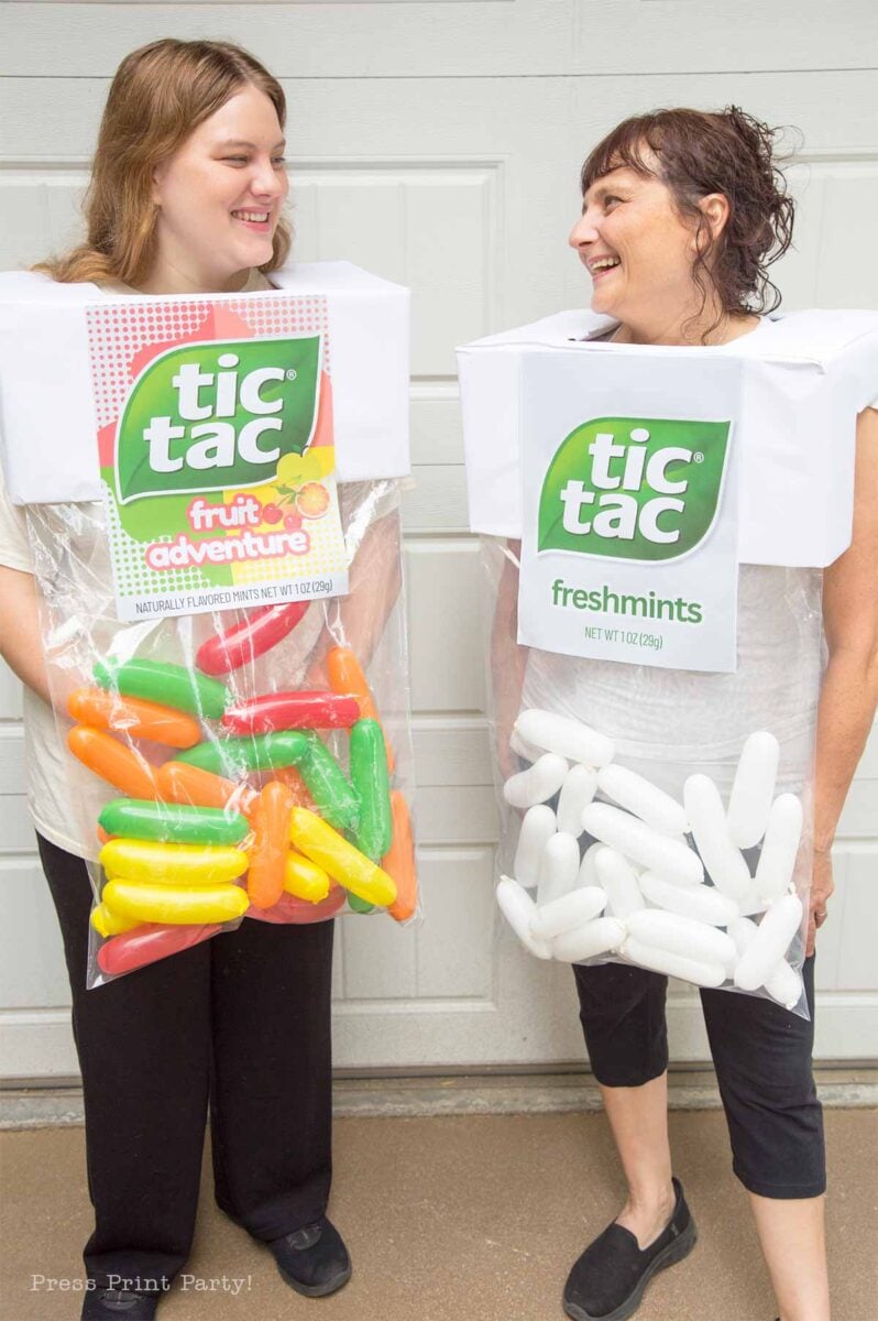 Two women wearing creative costumes made to look like Tic Tac candy boxes, one labeled "fruit adventure" with colorful candies and the other "freshmints" with white candies. Both are smiling and standing in front of a garage door.