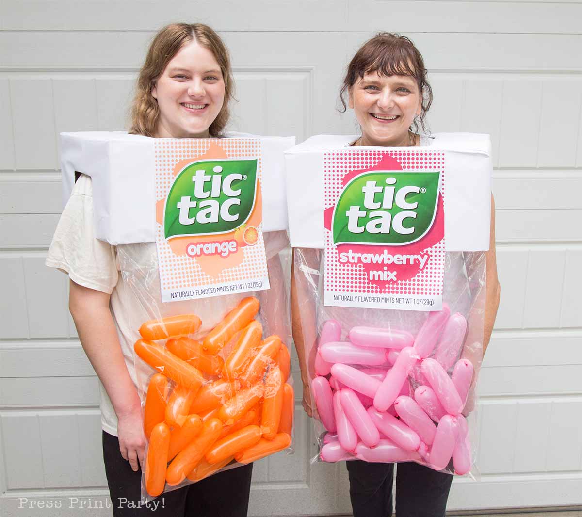 Two smiling women wearing homemade Tic Tac costumes stand in front of a garage. One costume is orange with orange balloons, labeled "orange," and the other is pink with pink balloons, labeled "strawberry mix.