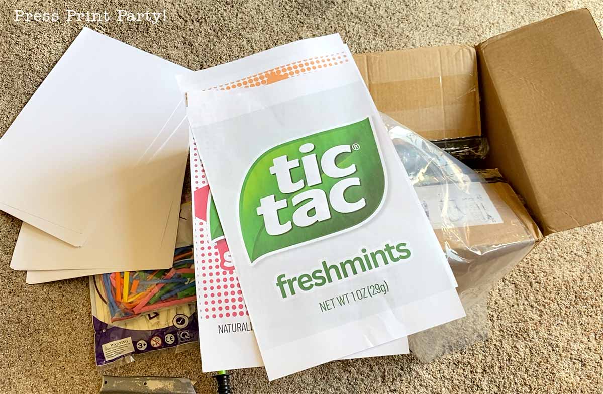 A large paper with a Tic Tac Freshmints label sits on top of a stack of papers, boxes, and colorful crafting supplies on a carpeted floor.