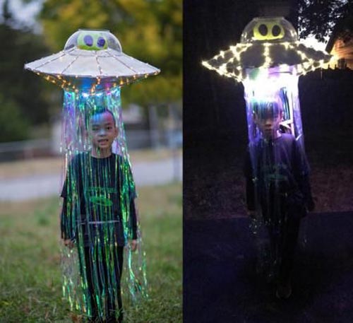 A child wears a UFO costume with a large, saucer-shaped hat decorated with lights and shiny streamers hanging down, creating the effect of a spaceship beaming them up. The costume is shown during the day and glowing at night.
