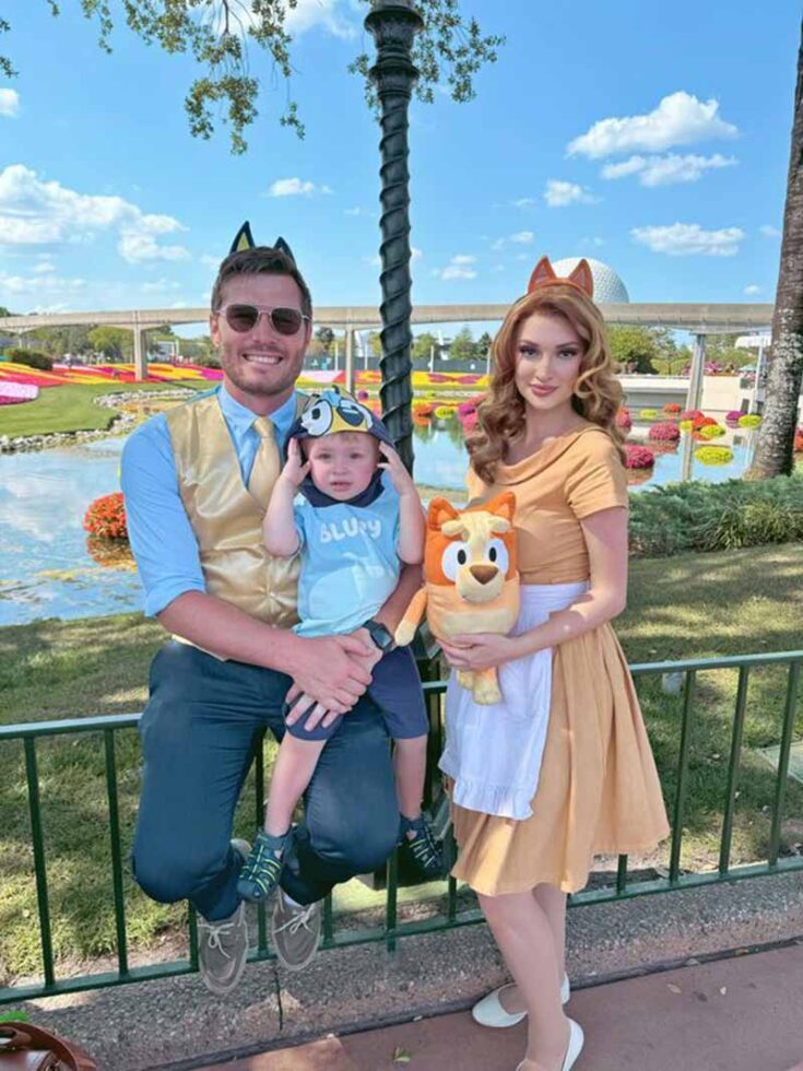 A family of three poses outdoors in front of a garden and pond, dressed in Bluey-themed costumes. Showcasing one of the cutest family costume ideas, they wear animal ear headbands and a Bluey cap, while the mother holds a Bluey plush toy.