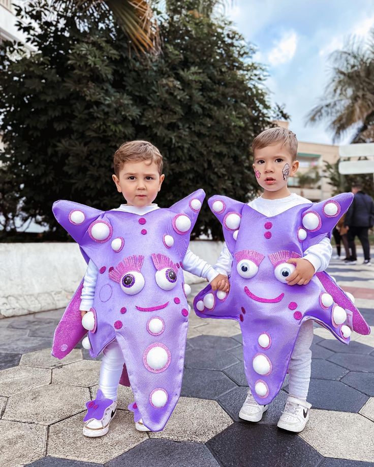 Two young children stand outdoors wearing matching purple starfish costumes with big eyes, pink eyelashes, and white spots. They are holding hands and have face paint on their cheeks. Trees and clouds are visible in the background.