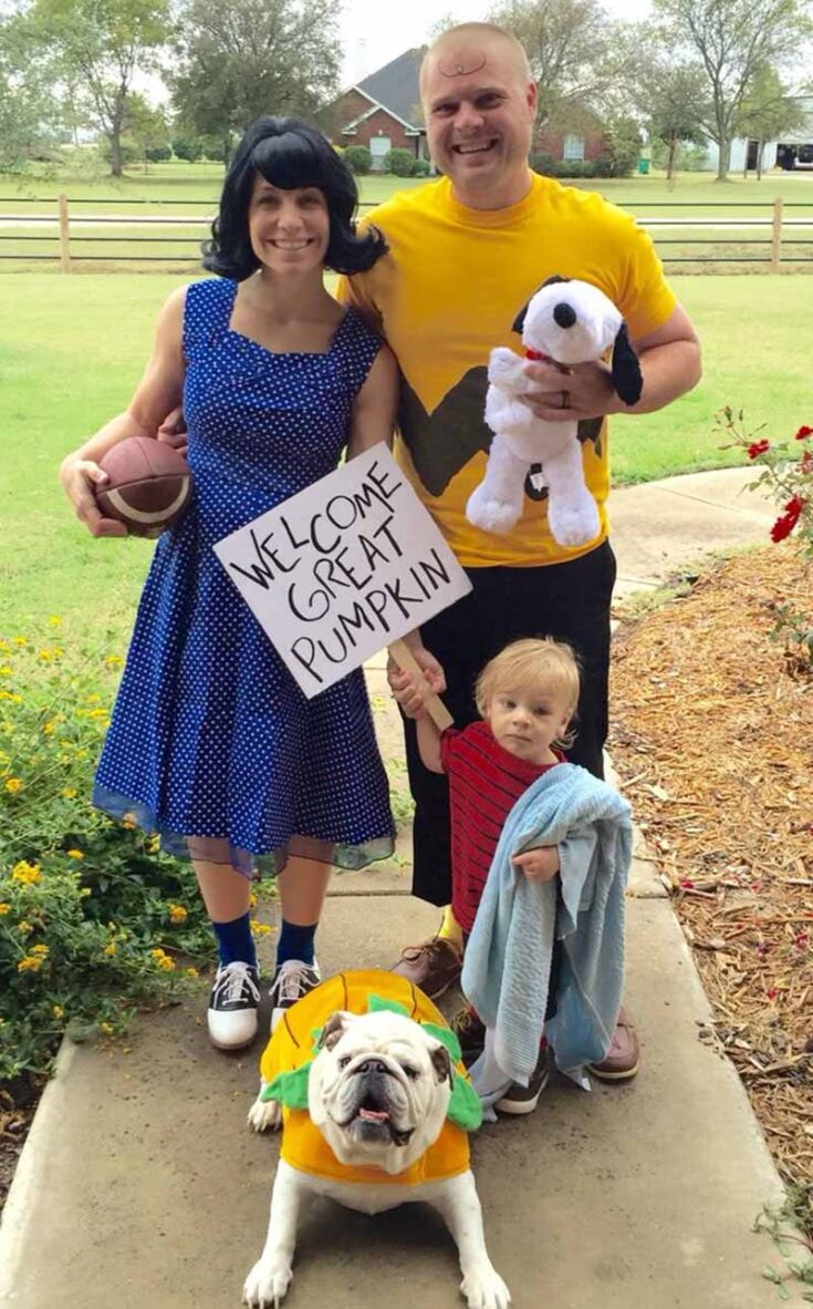 A family dressed as Peanuts characters—Lucy, Charlie Brown, Linus with a blue blanket, and a bulldog in a pumpkin costume—hold a “Welcome Great Pumpkin” sign. Perfect inspiration for creative family costume ideas.