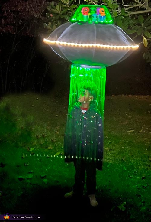 A child stands under a glowing UFO costume with green lights and hanging streamers, creating the effect of being beamed up by an alien spaceship at night.