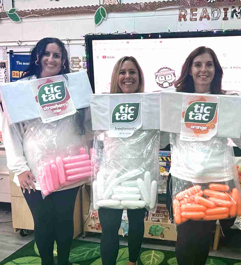 Three women dressed in creative Tic Tac costume boxes—one as strawberry (pink), one as freshmints (white), and one as orange flavor—stand smiling in a classroom with books and decorations in the background.
